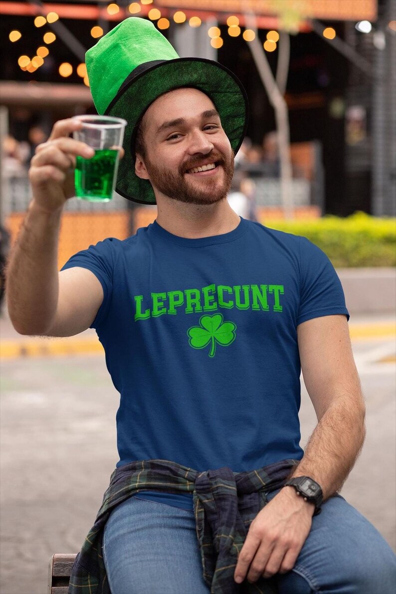 A man wearing a blue t-shirt with the text "Leprecht" and a green hat is sitting on a bench, holding a drink in his hand.