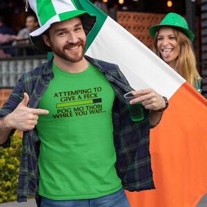 A man wearing a green t-shirt with a humorous text and a green hat is holding an orange flag, while a woman in a green hat is smiling behind him.