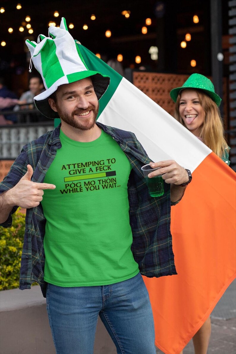 A man wearing a green t-shirt with a humorous text and a green hat is holding an orange flag, while a woman in a green hat is smiling behind him.