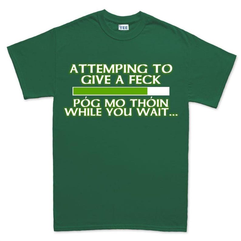The image is a green t-shirt with white text that reads "ATTEMPTING TO GIVE A FEK PÓG MO THÍON WHILE YOU WAIT...".