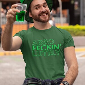 May include: A green t-shirt with the text "ABSO FECKIN' LUTELY" in green. The person in the image is wearing a green leprechaun hat and holding a clear plastic cup with green liquid. The person is smiling and wearing a watch.