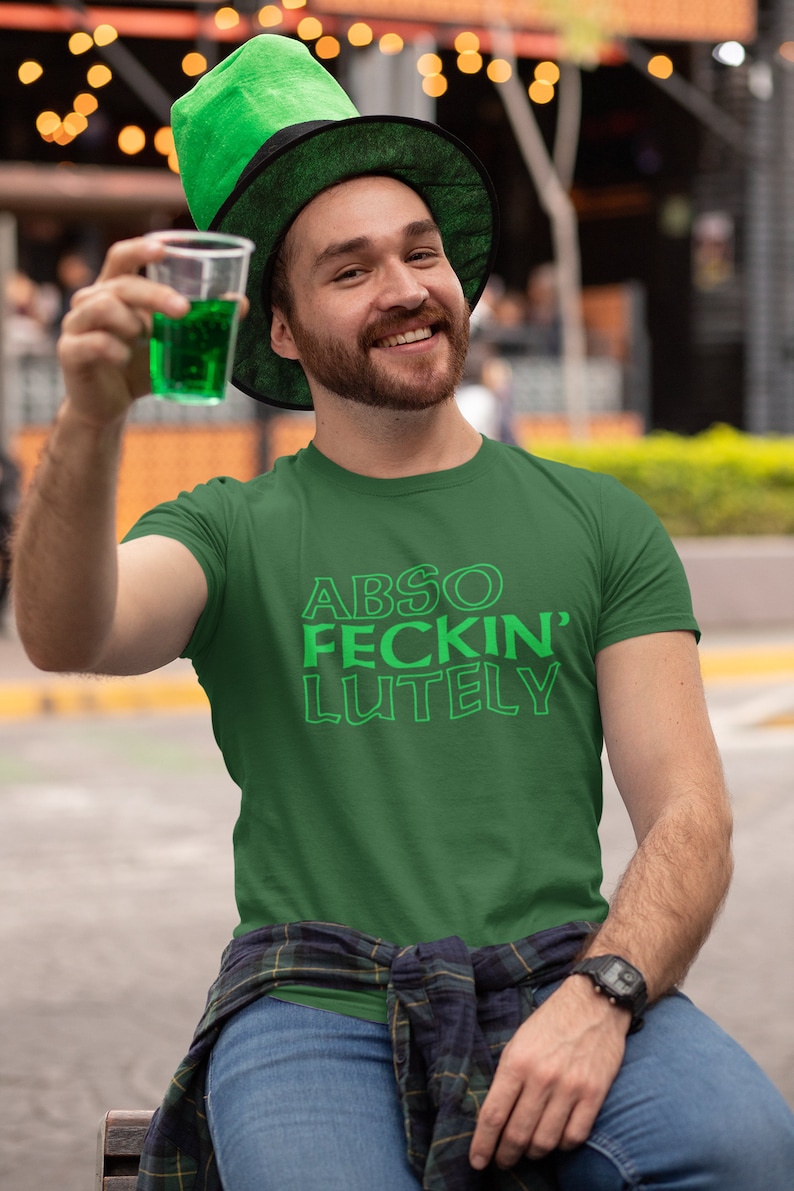 May include: A green t-shirt with the text "ABSO FECKIN' LUTELY" in green. The person in the image is wearing a green leprechaun hat and holding a clear plastic cup with green liquid. The person is smiling and wearing a watch.