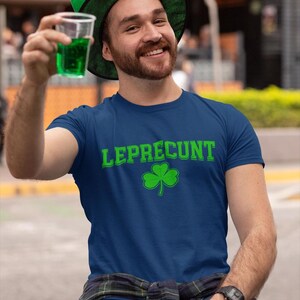 A man wearing a blue t-shirt with the text "Leprecht" and a green hat is sitting on a bench, holding a drink in his hand.