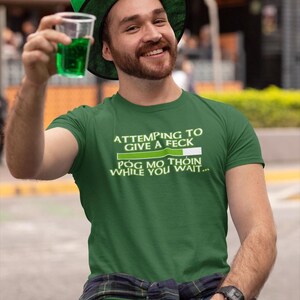 A man wearing a green hat and a green t-shirt is sitting on a bench, holding a drink and smiling.