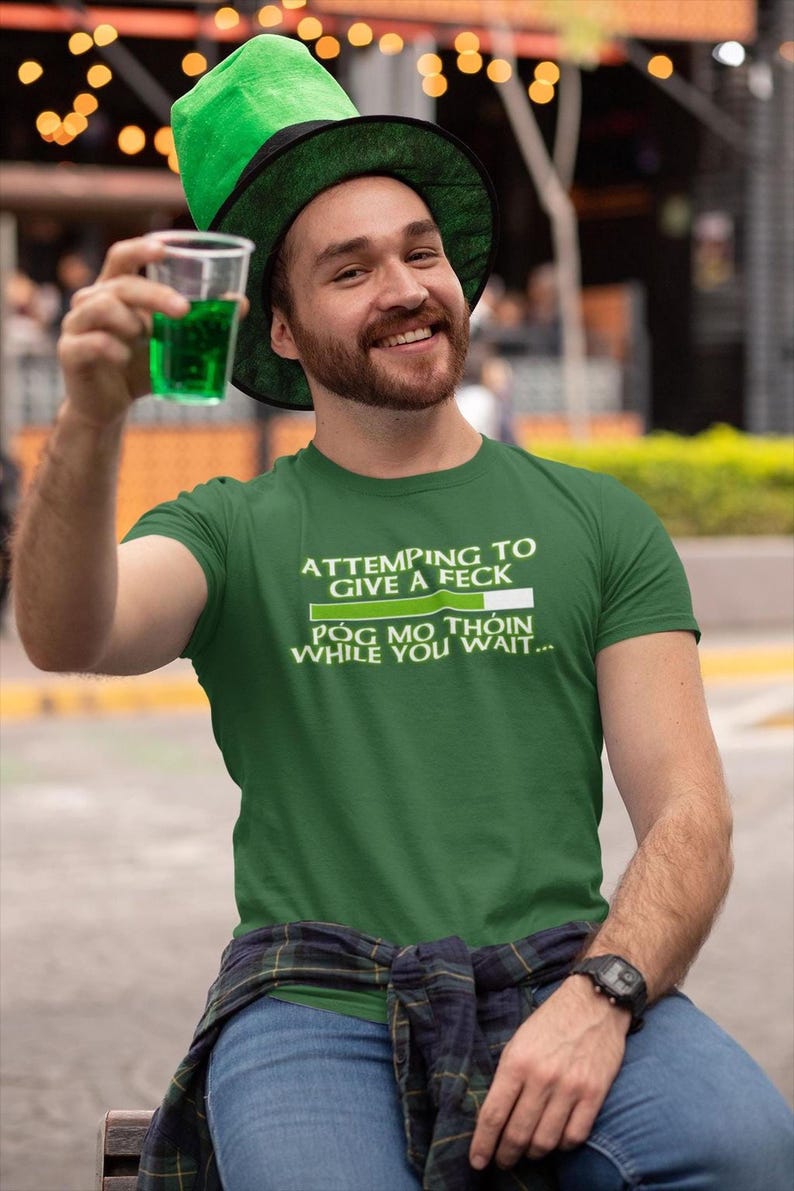 A man wearing a green hat and a green t-shirt is sitting on a bench, holding a drink and smiling.