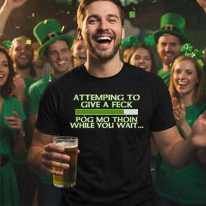 A man wearing a black t-shirt with a humorous text is smiling and holding a glass of beer, surrounded by a group of people wearing green hats and smiling.