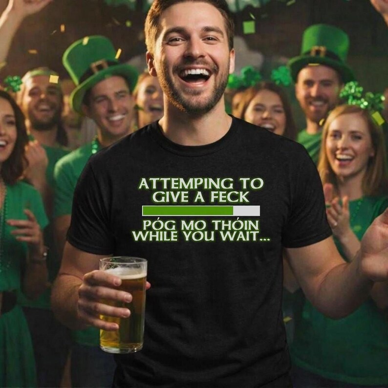 A man wearing a black t-shirt with a humorous text is smiling and holding a glass of beer, surrounded by a group of people wearing green hats and smiling.