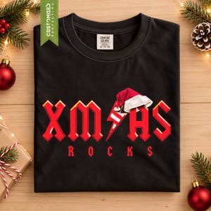 Peut inclure: Un t-shirt noir avec le texte rouge XMAS, où un éclair remplace la lettre A, surmonté d'un bonnet de Père Noël rouge et blanc. En dessous, le mot ROCKS est imprimé en rouge. Le t-shirt porte une étiquette Comfort Colors à l'encolure.
