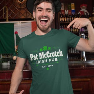 A man wearing a green hat and a green t-shirt with white text is smiling and pointing at the camera in a bar setting.