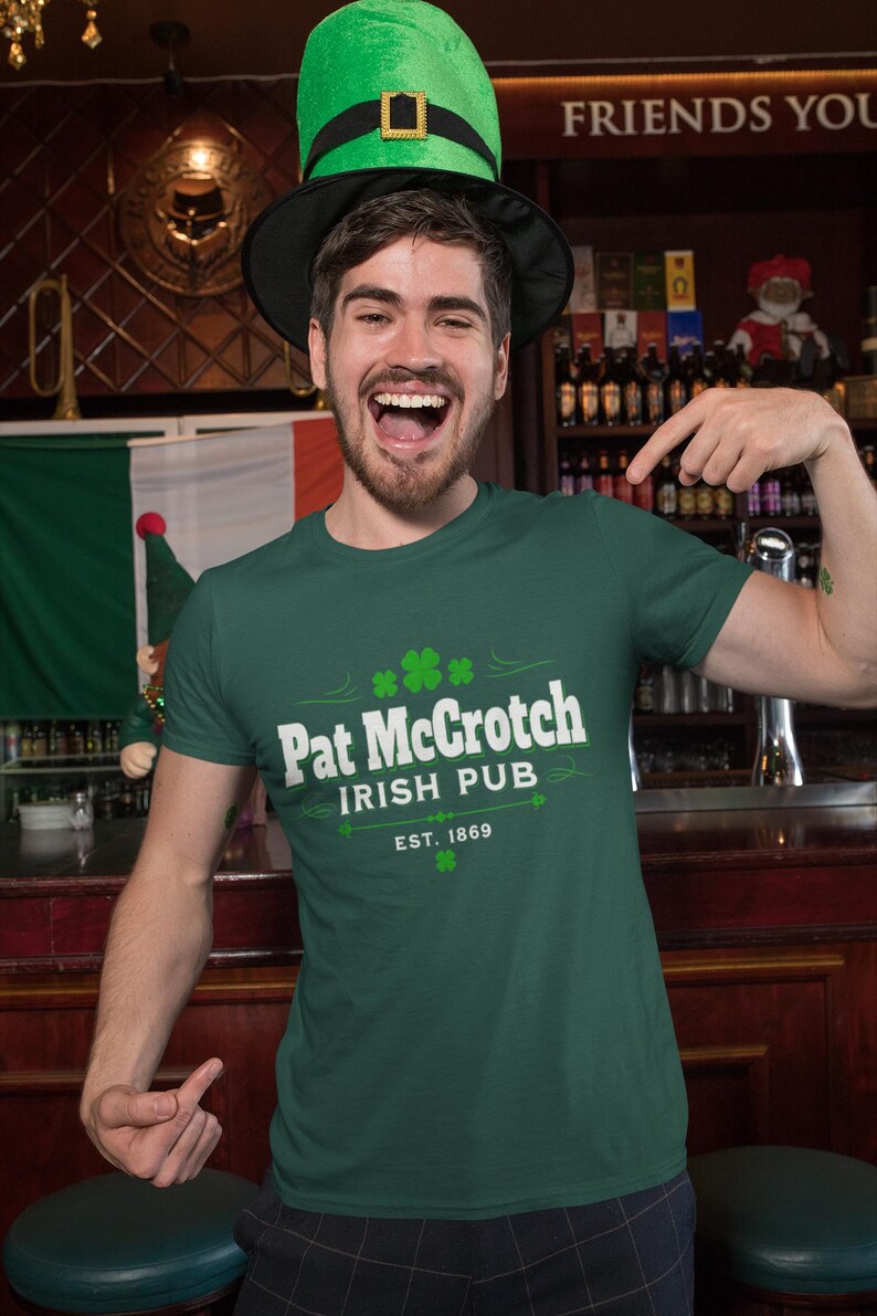 A man wearing a green hat and a green t-shirt with white text is smiling and pointing at the camera in a bar setting.