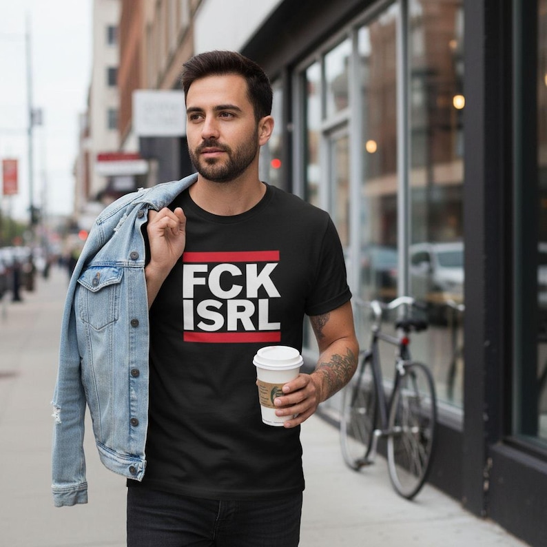 A man wearing a black t-shirt with the text "FCK ISRL" is walking down a city street while holding a coffee cup.