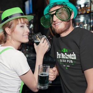 A woman wearing a green hat and a man wearing a green hat and a t-shirt with the text "Pat McC Scotch Irish Pub" are posing together, holding glasses of beer in a bar setting.