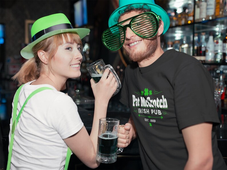 A woman wearing a green hat and a man wearing a green hat and a t-shirt with the text "Pat McC Scotch Irish Pub" are posing together, holding glasses of beer in a bar setting.