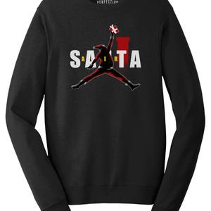 Könnte beinhalten: Ein schwarzes Langarm-Sweatshirt mit einer Grafik, die die Silhouette des Weihnachtsmanns beim Springen mit einem Geschenk zeigt. Der Text "SANTA" ist in weißen Buchstaben hinter der Figur gedruckt, mit einem gelben "I" und einer roten Kaminform.