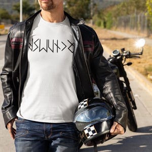 A man wearing a white t-shirt and a black leather jacket stands next to a motorcycle, holding a helmet.