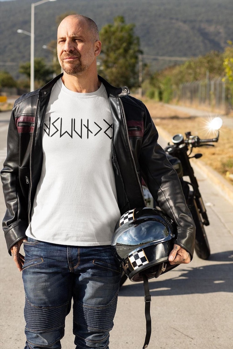 A man wearing a white t-shirt and a black leather jacket stands next to a motorcycle, holding a helmet.