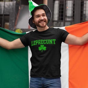 A man wearing a black t-shirt with the word "LEPRECIUNT" and a green hat is holding up an orange and green flag, smiling and posing for the camera.