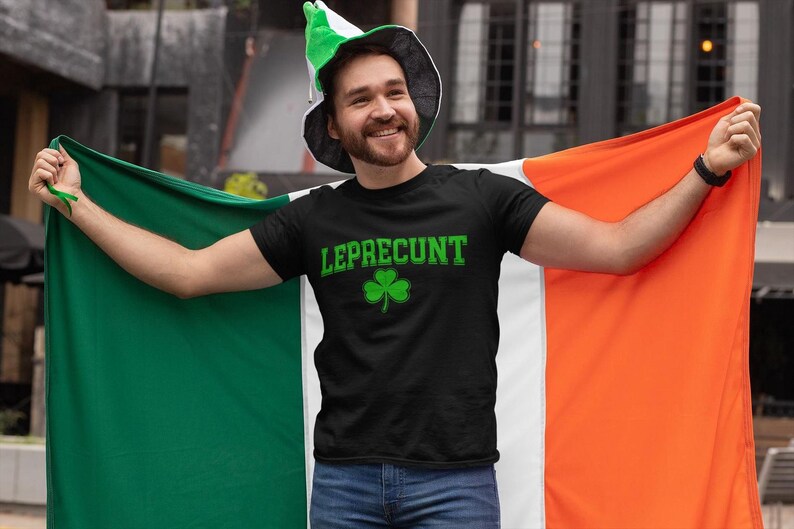 A man wearing a black t-shirt with the word "LEPRECIUNT" and a green hat is holding up an orange and green flag, smiling and posing for the camera.