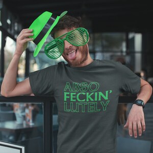 May include: Dark gray t-shirt with the text "ABSO FECKIN' LUTELY" in green. The person in the image is wearing green shutter shades and a green hat. The person is leaning on a glass railing.