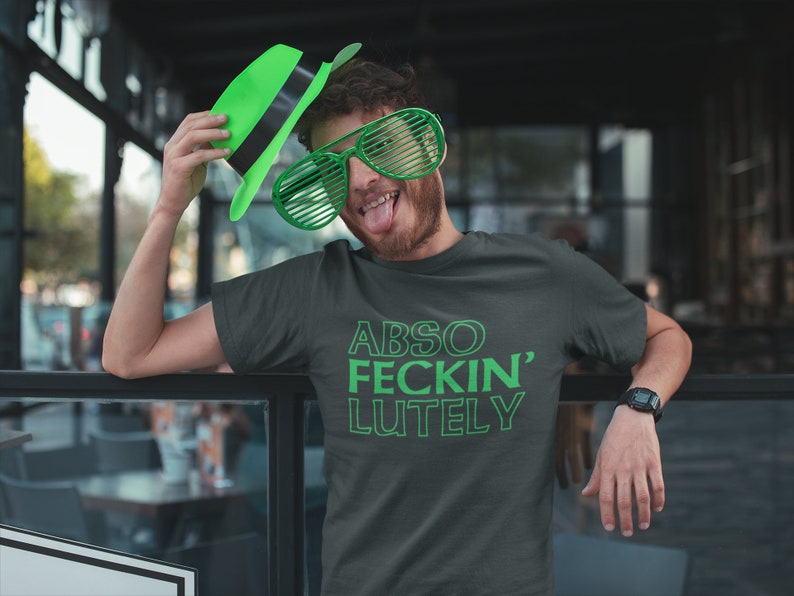 May include: Dark gray t-shirt with the text "ABSO FECKIN' LUTELY" in green. The person in the image is wearing green shutter shades and a green hat. The person is leaning on a glass railing.