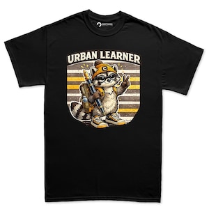 Op de afbeelding: Een zwart t-shirt met een afbeelding van een wasbeer die een gele muts, een bril en een rugzak draagt. De wasbeer houdt een grote vulpen vast en maakt een vredesteken. De tekst URBAN LEARNER staat boven de wasbeer op een gestreepte achtergrond.