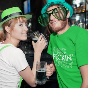 May include: A couple celebrating St. Patrick's Day in a bar. The woman wears a green hat and suspenders, and the man wears a green t-shirt with the text "ABSO FECKIN' LUTELY" and green shutter shades. Both hold mugs of green beer.