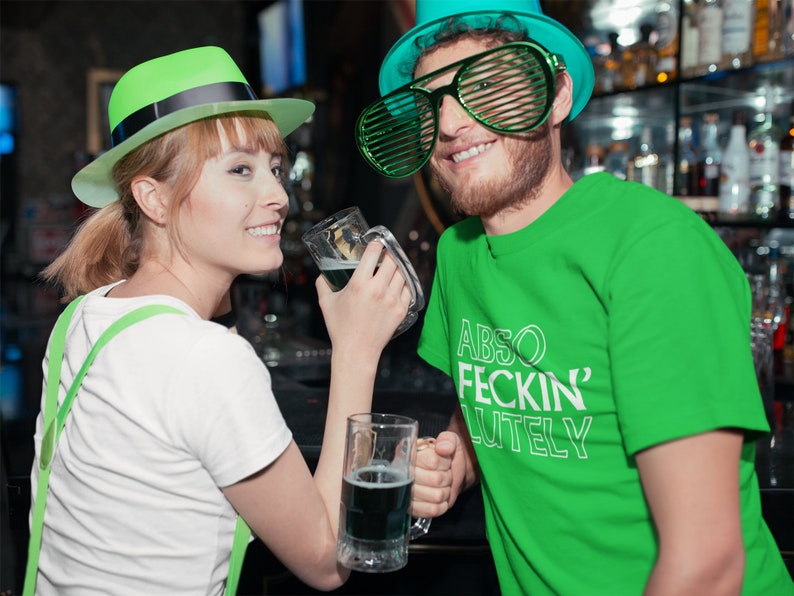 May include: A couple celebrating St. Patrick's Day in a bar. The woman wears a green hat and suspenders, and the man wears a green t-shirt with the text "ABSO FECKIN' LUTELY" and green shutter shades. Both hold mugs of green beer.