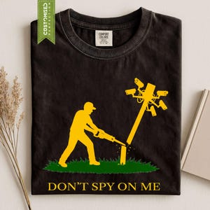 May include: A black crew neck t-shirt featuring a yellow graphic of a person using a chainsaw to cut down a utility pole topped with multiple security cameras. Below the graphic, the text reads DON'T SPY ON ME in yellow capital letters.