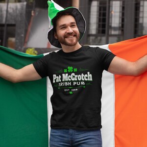 A man wearing a black t-shirt with the text "Pat McCroch Irish Pub EST 1818" and a green hat is holding up an orange and green flag, smiling and posing for the camera.