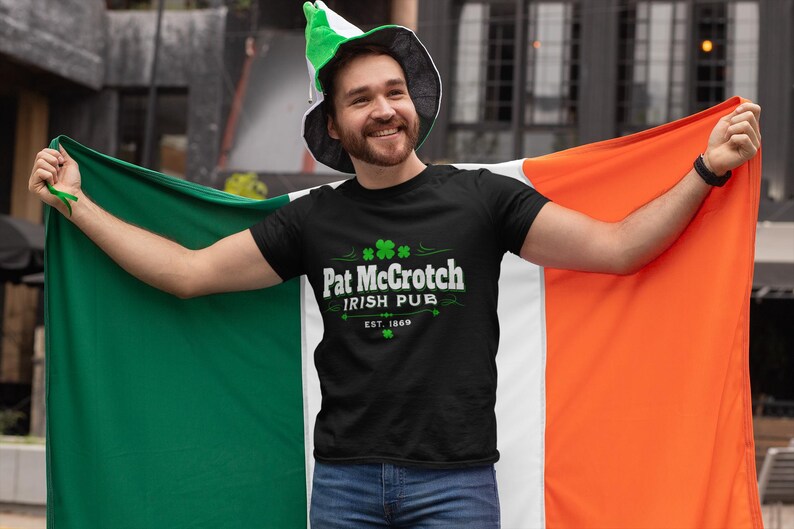 A man wearing a black t-shirt with the text "Pat McCroch Irish Pub EST 1818" and a green hat is holding up an orange and green flag, smiling and posing for the camera.