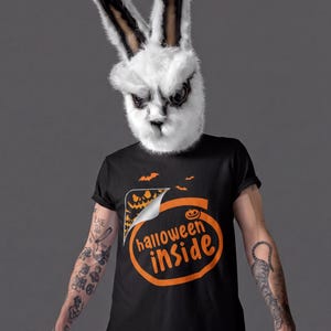 A person wearing a bunny mask and a Halloween-themed t-shirt stands against a gray background.