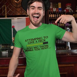 A man wearing a green t-shirt with a humorous text and a green leprechaun hat is standing in front of a bar.