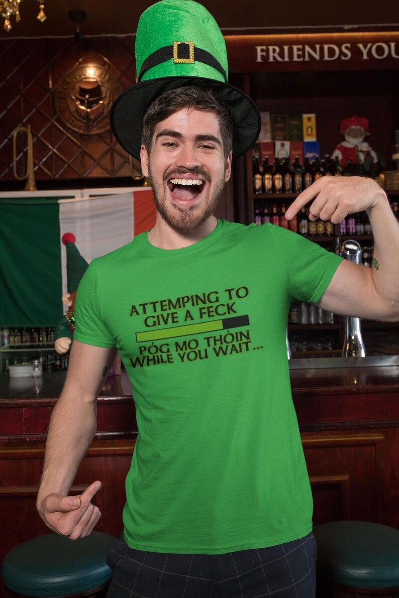 A man wearing a green t-shirt with a humorous text and a green leprechaun hat is standing in front of a bar.