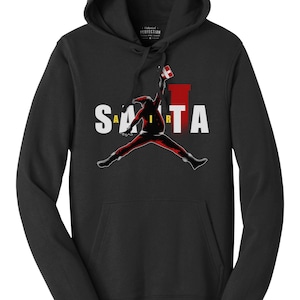 Könnte beinhalten: Schwarzer Kapuzenpullover mit einem Grafikdruck von Weihnachtsmann, der in Rot und Weiß gekleidet ist, mit einem Geschenk in der Hand durch die Luft springt. Der Text "SANTA AIR" ist in gelben Buchstaben auf dem Pullover gedruckt.