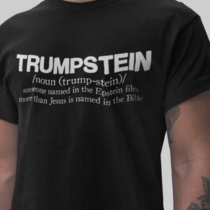 Può includere: T-shirt nera con la parola "TRUMPSTEIN" in grandi lettere bianche. Sotto, una definizione: "/sostantivo (trump-stein)/ qualcuno nominato nei file di Epstein più di Gesù nella Bibbia."