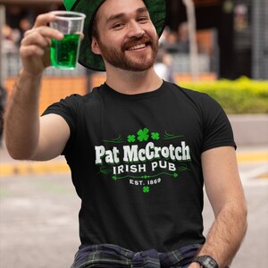 A man wearing a green hat and a black t-shirt with the text "Pat Crotch Irish Pub EST.1869" is sitting on a bench, holding a glass of green liquid and smiling.