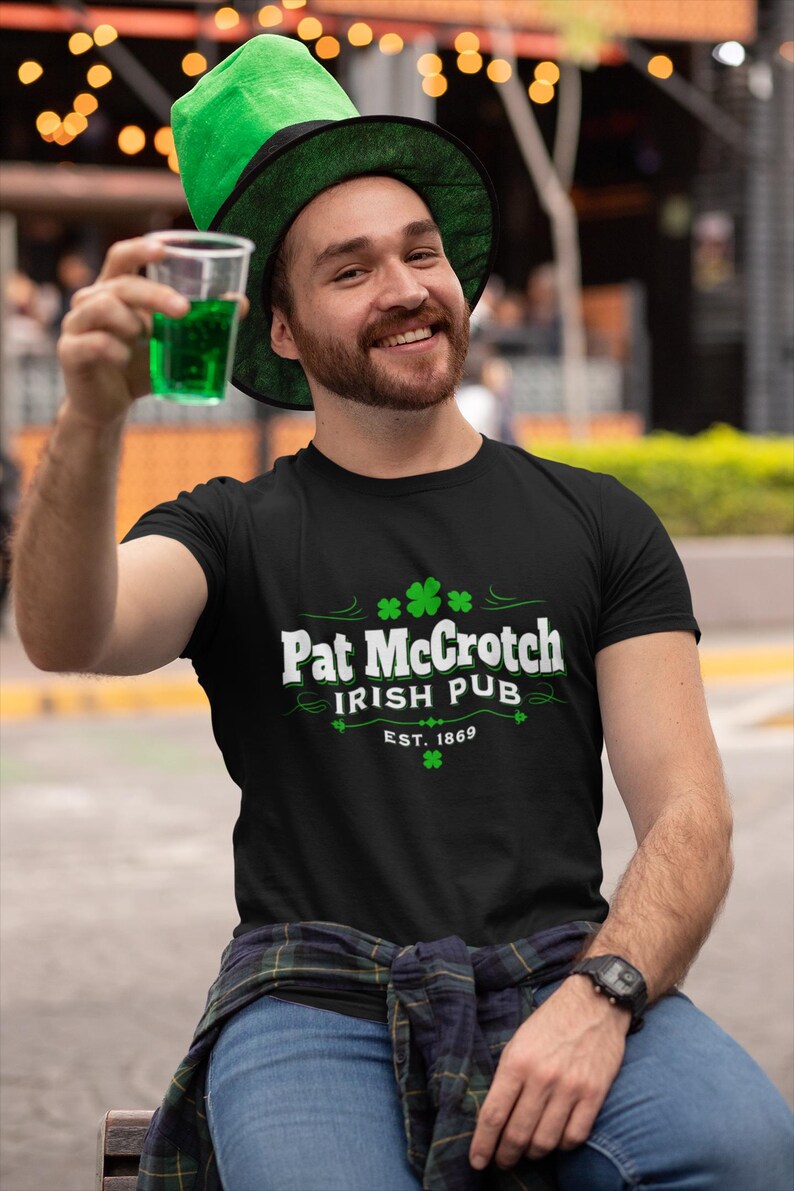 A man wearing a green hat and a black t-shirt with the text "Pat Crotch Irish Pub EST.1869" is sitting on a bench, holding a glass of green liquid and smiling.