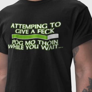A black t-shirt with white text that reads "ATTEMPTING TO GIVE A FEK POG MO THION WHILE YOU WAIT..."