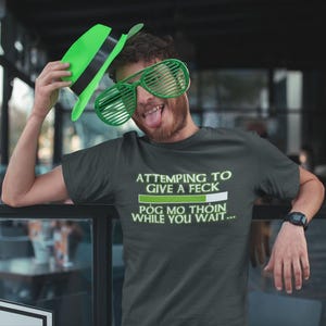A man wearing a green hat and large sunglasses is posing for a photo, with a t-shirt that says "ATTEMPTING TO GIVE A FECK POG MO THOIN WHILE YOU WAIT..."
