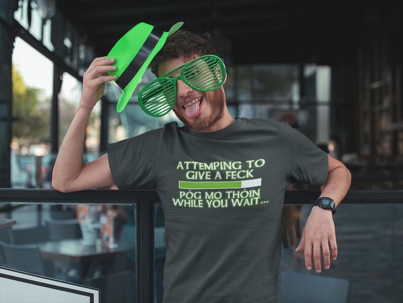A man wearing a green hat and large sunglasses is posing for a photo, with a t-shirt that says "ATTEMPTING TO GIVE A FECK POG MO THOIN WHILE YOU WAIT..."