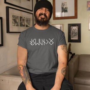 A man with a beard and a black beanie is sitting in a living room, wearing a gray t-shirt with white text and tattoos on his arms.