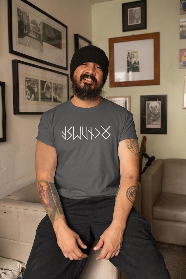 A man with a beard and a black beanie is sitting in a living room, wearing a gray t-shirt with white text and tattoos on his arms.