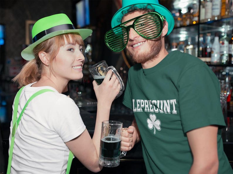 A woman wearing a green hat and a man wearing a green hat with large glasses are standing together, holding glasses of beer and smiling.