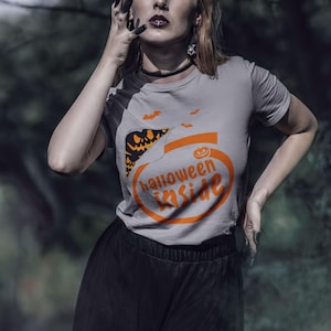 A woman wearing a Halloween-themed t-shirt stands in a forest, with a serious expression on her face.