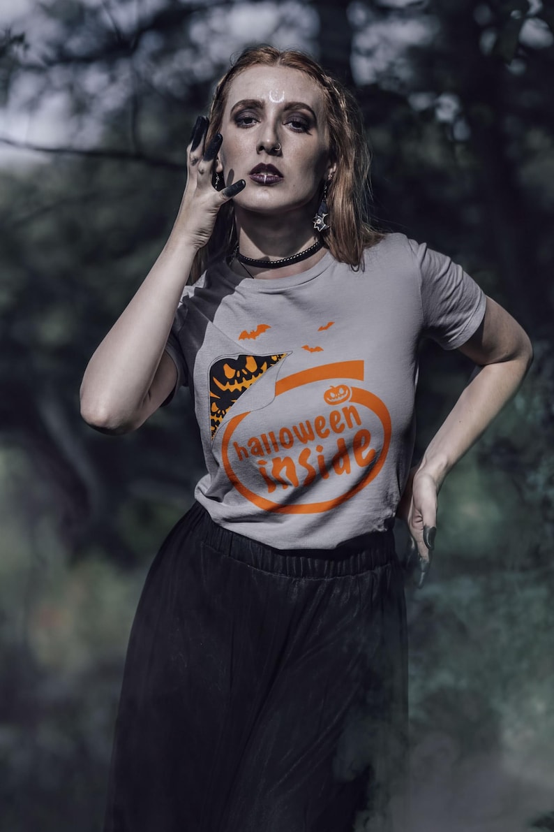A woman wearing a Halloween-themed t-shirt stands in a forest, with a serious expression on her face.