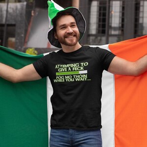 A man wearing a black t-shirt with a humorous Irish flag design and a green hat is holding up an orange and green flag, smiling and appearing to be in a celebratory mood.