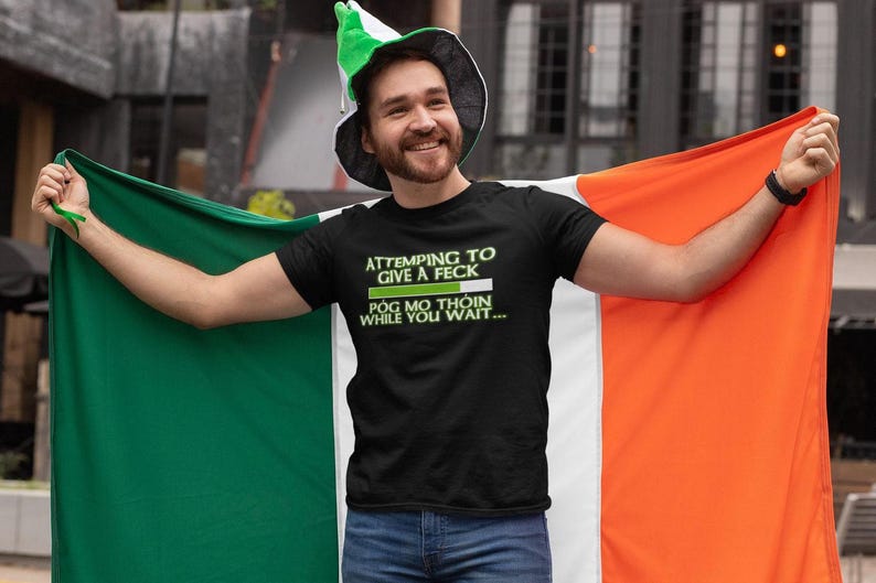 A man wearing a black t-shirt with a humorous Irish flag design and a green hat is holding up an orange and green flag, smiling and appearing to be in a celebratory mood.