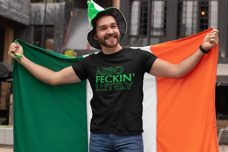 May include: Black t-shirt with the text "ABSO FECKIN' LUTELY" in green. The person is holding an Irish flag, which is green, white, and orange. The person is wearing a green and white hat.