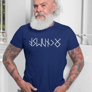 A man with a long white beard and tattoos on his arms is wearing a blue t-shirt with white text on it.