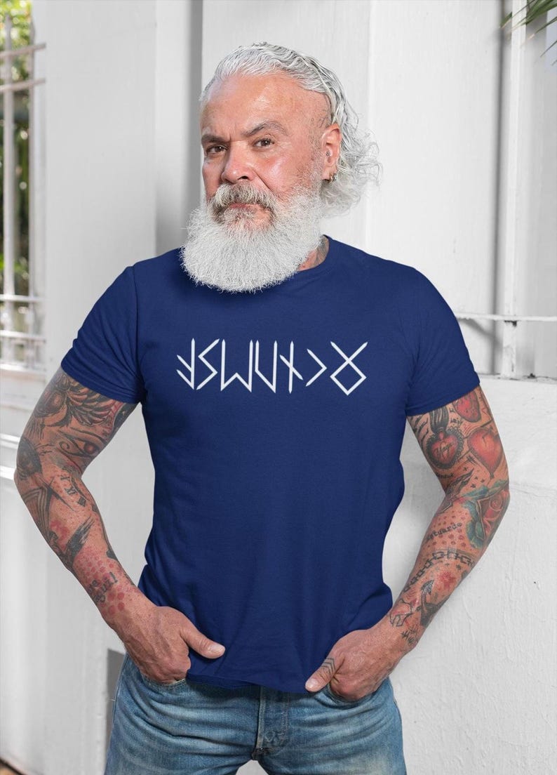 A man with a long white beard and tattoos on his arms is wearing a blue t-shirt with white text on it.